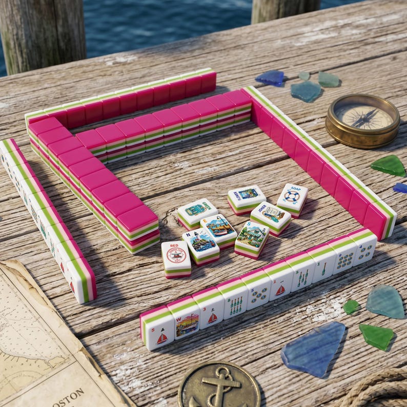 May include: A Mahjong set featuring tiles with pink and green edges, arranged in a partial square formation on a wooden surface. Several loose tiles display nautical illustrations, including lighthouses and sailboats, alongside a brass compass and anchor.