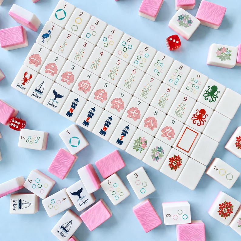 May include: A complete Mahjong set with white tiles featuring various symbols and numbers, including jokers, lighthouses, and floral designs. The set includes pink tile holders and a red die, all arranged on a light blue surface.
