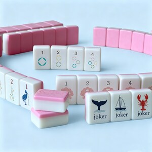 May include: A set of pink and white Mahjong tiles arranged in a circular pattern. The tiles feature various symbols, numbers, and illustrations, including a whale tail, sailboat, and lobster. The game pieces are set against a light blue background.