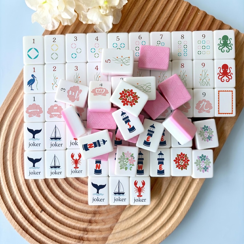 May include: A collection of white and pink Mahjong tiles with various designs, including numbers, flowers, and nautical themes. The tiles are arranged on a wooden tray, with some pink tiles scattered around.