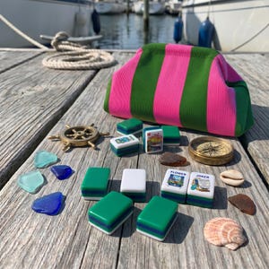 May include: A fabric pouch with vertical pink and green stripes, accompanied by a collection of mahjong tiles, a small brass ship wheel, a brass compass, several pieces of colorful sea glass, and two seashells arranged on a wooden surface.