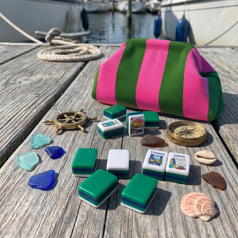 May include: A fabric pouch with vertical pink and green stripes, accompanied by a collection of mahjong tiles, a small brass ship wheel, a brass compass, several pieces of colorful sea glass, and two seashells arranged on a wooden surface.