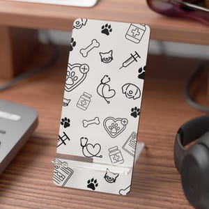 Può includere: Un supporto per telefono bianco con un motivo nero ripetuto di icone relative agli animali domestici, tra cui impronte di zampe, ossa, facce di gatto e simboli medici. Il supporto è progettato per tenere un telefono in posizione verticale.