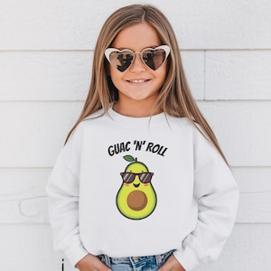 Può includere: Felpa bianca con la scritta "GUAC 'N' ROLL" e un avocado a cartoni animati con occhiali da sole. L'avocado è verde e marrone. La felpa è indossata da una persona con occhiali da sole a forma di cuore.