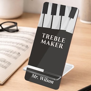 May include: A black acrylic phone stand with a piano key design at the top and the words "TREBLE MAKER" in white. The name "Mr. Wilson" is printed at the bottom. The stand is on a wooden surface with sheet music and glasses in the background.