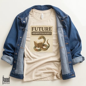 Può includere: Una maglietta color crema con una grafica di un serpente che legge un libro, con le parole "FUTURE HERPETOLOGIST" sopra. La maglietta è sotto una giacca di jeans blu.