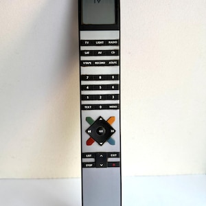 Pode incluir: Um controle remoto Bang & Olufsen prateado e preto com um design retangular. O controle remoto possui botões para TV, luz, rádio e outras funções. A seção superior exibe "TV". A parte inferior do controle remoto tem o nome da marca.