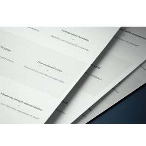 May include: Close-up of several white paper sheets with printed text. The text includes phrases like "I create space for peace" and "I am anchored in love." The sheets are slightly fanned out, revealing the text on each page. The background is a dark blue.