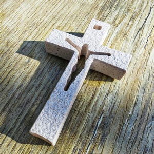 Puede incluir: Una cruz de piedra tallada con una superficie texturizada. La cruz presenta un diseño recortado con una figura en el centro. La cruz es de color gris claro y está sobre una superficie de madera.