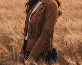 May include: A brown suede jacket with fringe detailing on the sleeves and chest. The jacket has a classic collar and is worn over a white top. The model is standing in a field of wheat.
