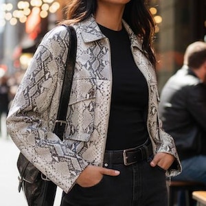 Pode incluir: Uma mulher usando uma jaqueta com estampa de pele de cobra, top preto e jeans preto. A jaqueta é cinza claro e branca com botões e dois bolsos no peito. Ela está usando um cinto preto e carregando uma bolsa preta.