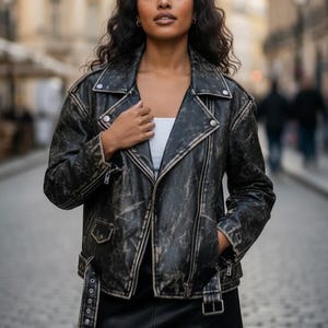 May include: A black distressed leather jacket with a biker style, featuring a notched collar, an asymmetrical zip, and pockets. The jacket has a worn, vintage appearance, with a belt detail at the waist. The model is wearing a white top and a black skirt.