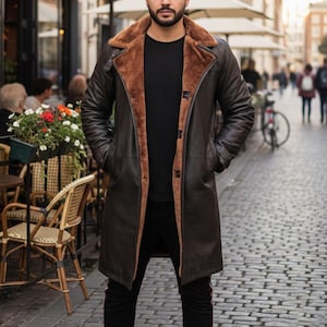 May include: A dark brown leather coat with a shearling collar and lining. The long coat has a button closure and pockets. The person in the image is wearing a black shirt and black trousers. The coat appears to be a winter coat.