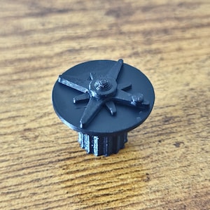 Puede incluir: Un pomo de plástico negro con una parte superior circular, un diseño en forma de estrella en relieve y una protuberancia central. La base del pomo es cilíndrica con estrías verticales en los lados, diseñada para un agarre funcional.