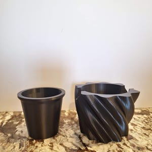 May include: A black, smooth-walled, tapered cup sits beside a black, cylindrical container featuring a deep, spiraling, vertical ribbed texture on its exterior. Both objects are made of a dark, solid material with a consistent matte finish.
