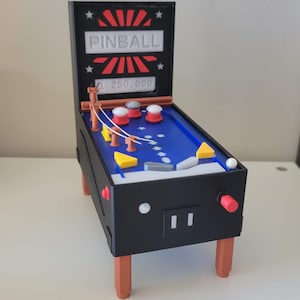 May include: A black tabletop pinball machine with orange legs, a blue playfield, and red and white bumpers. The backbox features the text PINBALL and a score of 1,250,000. It includes yellow triangular targets, a white ball, and a red plunger.