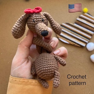 以下が含まれることがあります： 赤いリボンをつけた茶色の手編みの犬。犬は長い体と垂れた耳をしています。画像には、茶色と白のストライプが入ったハート型のアイテムと「Crochet pattern」というテキストも含まれています。