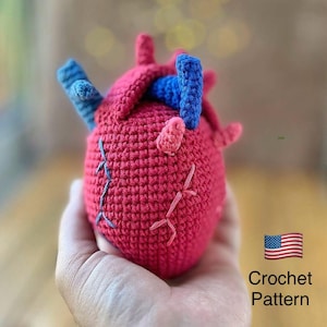 May include: A handmade crochet heart in shades of red, blue, and pink. The anatomical heart features blue and pink accents, with visible stitching details. The text "Crochet Pattern" is displayed next to the American flag.