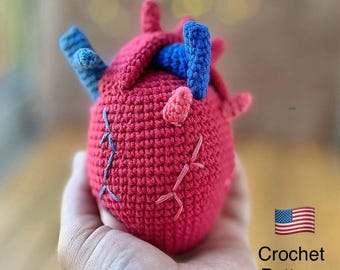 May include: A handmade crochet heart in shades of red, blue, and pink. The anatomical heart features blue and pink accents, with visible stitching details. The text "Crochet Pattern" is displayed next to the American flag.