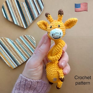 May include: A handmade, yellow crochet giraffe with brown accents. The giraffe has a white face and is held in a person's hand. The image also includes two heart-shaped decorations and the text "Crochet pattern".