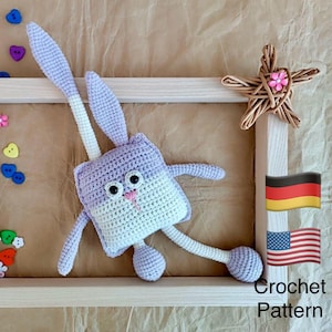 May include: A handmade crochet bunny toy in shades of lavender and white, with long ears and a square body. The bunny has black eyes and a pink nose. The image also includes a wooden frame, a woven star, and the text "Crochet Pattern".