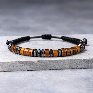 May include: A bracelet with alternating tiger's eye and black hematite beads. The bracelet has a black woven cord and is displayed on a grey surface. The tiger's eye beads are a warm, golden-brown colour. The bracelet is adjustable.