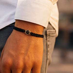 Könnte beinhalten: Schwarz geflochtenes Armband mit goldfarbenem Verschluss und einem dunklen, runden Stein. Das Armband wird am Handgelenk getragen. Ein weißes Hemd und Jeans sind im Hintergrund zu sehen. Ein stilvolles Accessoire.