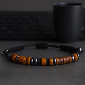 May include: A black braided bracelet with alternating brown and dark grey beads. The bracelet features a sliding knot closure for adjustable sizing. The beads are made of wood and hematite.