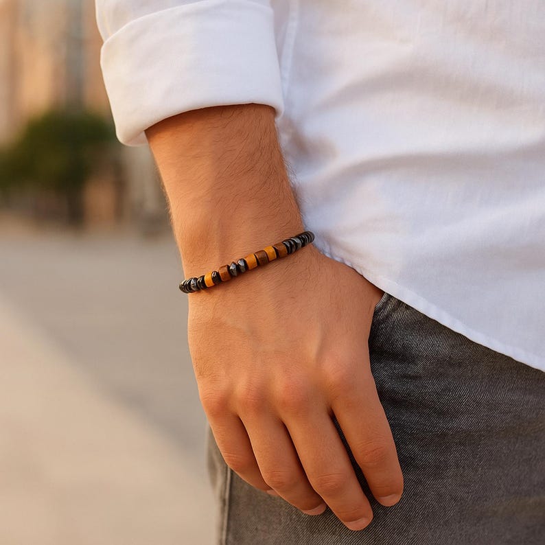 May include: A bracelet with alternating dark and amber-coloured beads. The bracelet is worn on a wrist, with a white shirt and grey trousers in the background. The design includes a mix of round and rectangular beads.
