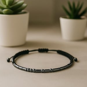 Può includere: Braccialetto di perline nere con cordino nero regolabile. Il braccialetto presenta una serie di piccole perline cilindriche e sferiche grigio scuro. Il braccialetto è esposto su una superficie neutra, con due piccole piante in vaso sullo sfondo.