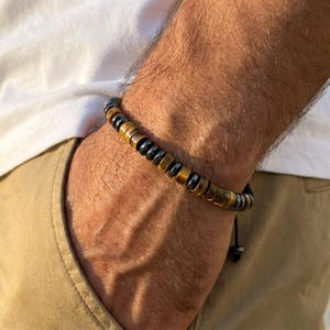 Pode incluir: Uma pulseira de contas com contas cilíndricas castanhas e pretas alternadas. A pulseira é usada no pulso, com uma camisa branca e calças cáqui no fundo. A pulseira é enfiada num cordão preto.