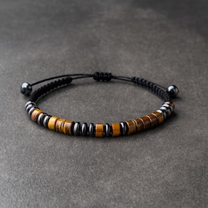 Puede incluir: Pulsera ajustable con cuentas de ojo de tigre y hematita. La pulsera tiene un cordón negro tejido con un cierre de nudo corredizo y dos pequeñas cuentas de color gris oscuro en los extremos. Las cuentas están dispuestas en un patrón repetitivo.