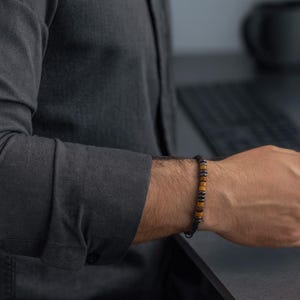 May include: A bracelet featuring brown, black, and gold beads. The bracelet is worn on a wrist, with the person wearing a dark grey long-sleeved shirt. The bracelet's design suggests it's made from natural materials, possibly wood or stone.