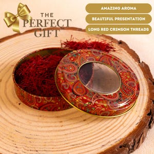 Puede incluir: Una lata redonda roja con un diseño de cachemira, llena de largos hilos carmesí. La tapa tiene una ventana transparente. El texto "THE PERFECT GIFT" es visible. La lata está sobre una superficie de madera.