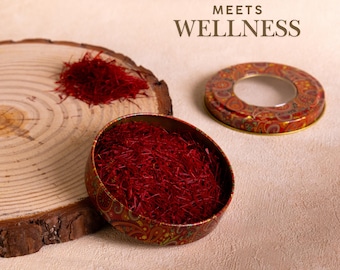 May include: A still-life image featuring saffron threads, a spice, presented on a wooden slice and in a decorative tin. The tin has a paisley design and a clear window. The text "TRADITION MEETS WELLNESS" is at the top of the image.