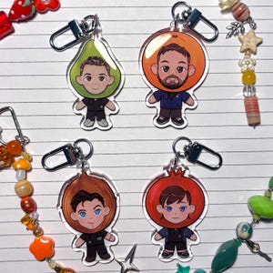 May include: Four clear acrylic keychains with cartoon characters in fruit costumes: a pear, orange, onion, and pomegranate. Each keychain has a silver clasp. The characters are dressed in dark uniforms.