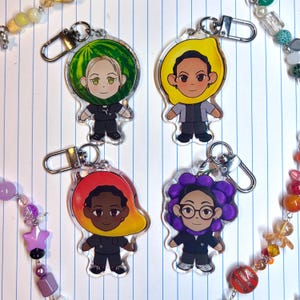 Puede incluir: Cuatro llaveros de acrílico con personajes de dibujos animados que llevan tocados con forma de fruta: sandía, limón, mango y un racimo de uvas. Cada personaje viste un uniforme oscuro y tiene un cierre de mosquetón de metal plateado.