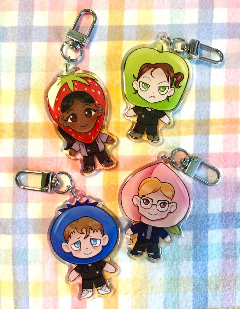 May include: Four acrylic keychains featuring cartoon characters in fruit-themed costumes: a strawberry, an apple, a blueberry, and a peach. Each character wears a black uniform and has a silver clasp for attachment.