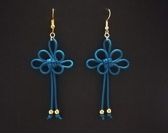 May include: A pair of handmade blue earrings with a floral knot design. Each earring has a gold-coloured hook and small gold beads at the bottom of the dangling cords. The earrings are set against a dark background.