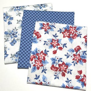 May include: Three folded fabric squares featuring distinct patterns. One square has a white background with blue and gray floral prints, one has a blue and white gingham check pattern, and one has a white background with red and blue floral prints.