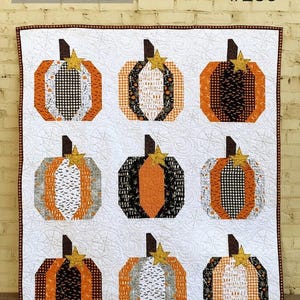 May include: A quilt featuring nine pumpkin blocks arranged in a grid on a white background with swirling patterns. Each pumpkin is constructed from various orange, black, white, and gray patterned fabrics with a small gold star accent.