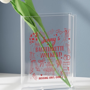 May include: A clear, rectangular vase with a red graphic design and text that reads "Jenny's Bachelorette Weekend" with the date December 12-13, 2025, and "Hocking Hills, Ohio". Two white tulips with green stems are inside the vase.