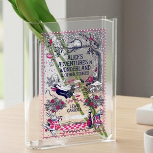 May include: A clear acrylic vase with a decorative panel featuring illustrations from "Alice's Adventures in Wonderland & Other Stories" by Lewis Carroll. The panel has a pink and white border with images of Alice, the White Rabbit, and other characters. White tulips are inside the vase.