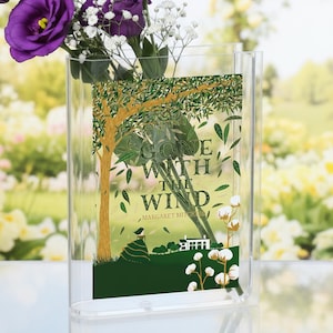 May include: A clear acrylic vase with a bouquet of purple flowers and baby's breath. The vase displays a book cover design with the title "Gone With The Wind" and a tree illustration. The background is a blurred garden scene.