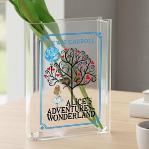May include: Clear acrylic vase with a design from "Alice's Adventures in Wonderland." The design features a tree with red heart-shaped leaves, a cat, and Alice. The vase holds white tulips. Text includes "Lewis Carroll" and "Alice's Adventures in Wonderland."