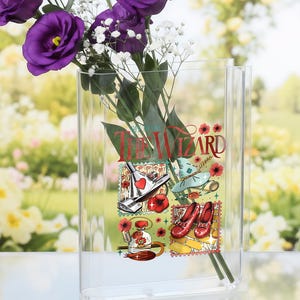 Op de afbeelding: Een heldere rechthoekige vaas met een bloemenontwerp met de tekst "The Wizard" en illustraties van een hart, schoenen en een hoed. De vaas bevat paarse bloemen en witte gipskruid.