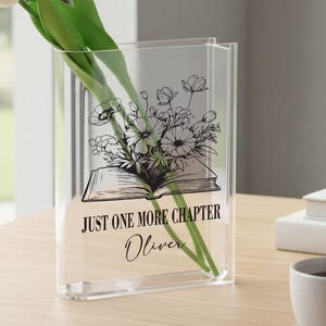 May include: A clear acrylic vase featuring a floral design and the text "JUST ONE MORE CHAPTER Oliver". The vase contains white tulips with green stems. The vase sits on a wooden table, next to a cup of coffee.