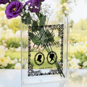 Op de afbeelding: Een heldere, rechthoekige vaas met paarse bloemen en witte gipskruid. De vaas is versierd met de tekst "Pride and Prejudice" en silhouetten van twee figuren, met daaronder "Jane Austen".