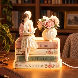 May include: A decorative lamp featuring a figurine of a person reading a book while sitting on a stack of four books. The books are labeled Emma, Just a Girl, and Who Loves Books. A small vase with pink roses sits on the top book.