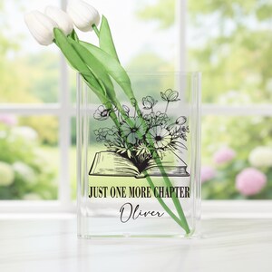May include: Clear acrylic vase with a floral design and the text "JUST ONE MORE CHAPTER Oliver." The vase holds white tulips with green stems and leaves. The floral design features an open book with flowers.